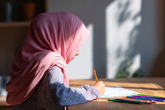 A schoolgirl wearing a pink hijab is focused on drawing with colored pencils at a desk
