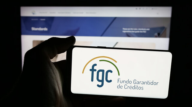 Stuttgart, Germany - 11-28-2025: Person holding mobile phone with logo of Brazilian Fundo Garantidor de Creditos (FGC) on screen in front of web page. Focus on phone display.