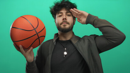 Young man with beard holding basketball makes salute gesture against green background showing...