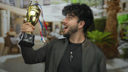 Young man with beard proudly holding a trophy outdoors on a sunny street with greenery around, embodying success and joy in an urban setting.