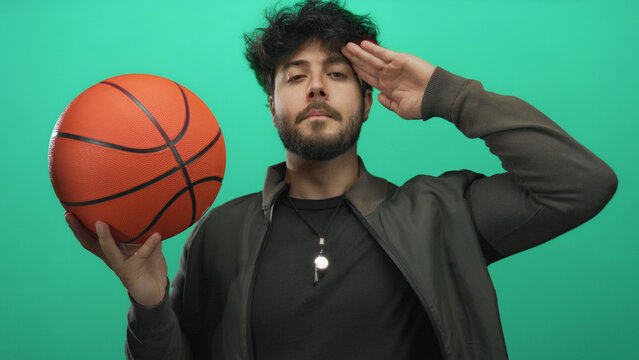 Young man with beard holding basketball makes salute gesture against green background showing sporty confident attitude - Powered by Adobe