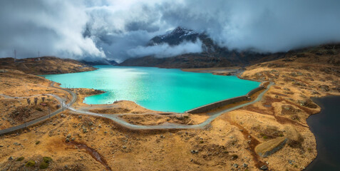 Fototapeta premium Aerial view of turquoise lake Bianco in the Swiss Alps, rocks and dramatic cloudy sky in autumn in Switzerland. Top drone view of alpine mountains, lake, trail, railroad, dam, orange hills in fall