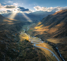 Fototapeta premium Aerial view of winding mountain road in the Swiss Alps, Switzerland, with sharp curves, golden slopes, valley and dramatic sun rays breaking through clouds over alpine peaks. Top drone view of road