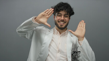 Fototapeta premium Young hispanic man with beard in glasses smiles and gestures in a white coat against a gray background displaying confidence and friendliness.