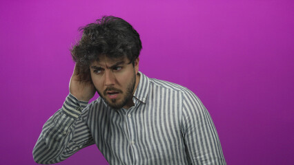 Young man with beard and striped shirt listens intently with hand to ear against vibrant pink background, conveying curiosity and focus in a modern setting.