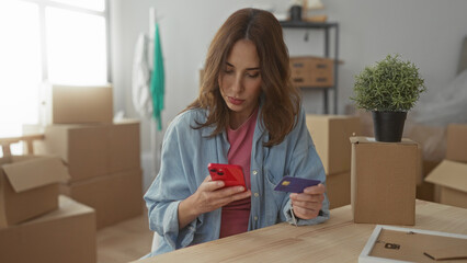 Woman using smartphone and credit card in new apartment surrounded by cardboard boxes plants and...