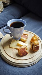 A cup of black coffee with sliced cheese and pastries arranged on a wooden board creates a cozy snack setting. The scene conveys comfort, relaxation and simple home lifestyle