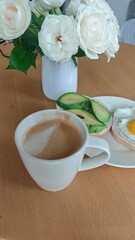 A cup of coffee is served beside a plate with avocado toast and a fried egg on a wooden table near a vase of white roses. The scene conveys a calm homemade breakfast and a fresh, peaceful morning mood