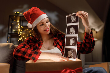 A woman wearing a Santa hat opens a gift box to reveal an ultrasound, celebrating a Christmas pregnancy announcement