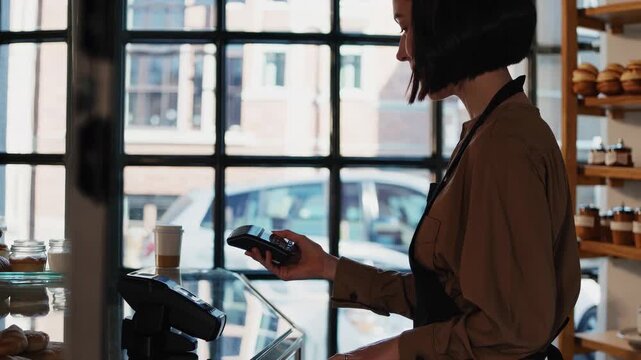 Cashier in bakery holds credit card terminal for payment. Customer makes contactless payment. Female worker at counter processes sale. Bakery service with modern payment. Small business and retail con