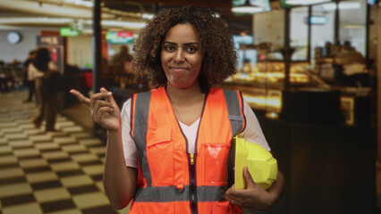 Woman engineer in orange safety vest holding yellow hardhat and pointing finger inside building; safety confidence.