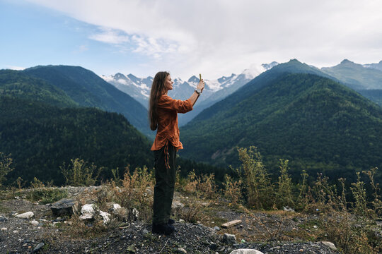 Woman stands on rocky ground in a dramatic mountain landscape, holding a device to take a photo, wearing an orange top, with pine trees and distant peaks