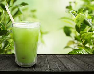 Matcha drink with ice in glass on wooden table against green leaves. Space for text