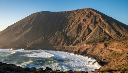 Large rugged coastal mountain with a winding road at its base is illuminated by golden sunlight as powerful ocean waves crash on the rocky shore below in a scenic landscape