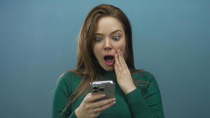 Young caucasian woman holding smartphone and mouth open in blue studio space touching cheek with expression of shock; surprise curiosity interest discovery.