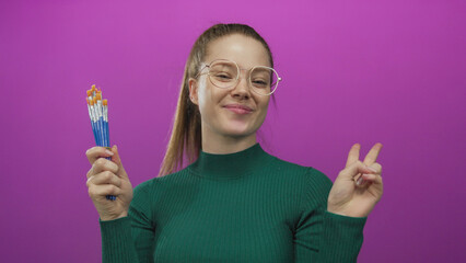 Woman holds several paintbrushes in studio setting while smiling with glasses and green turtleneck; creative joy.