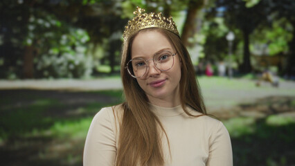 Young caucasian princess wearing a golden crown and thin glasses stands with crossed arms among blurred green foliage in a sunlit forest clearing  rejection. © Krakenimages.com