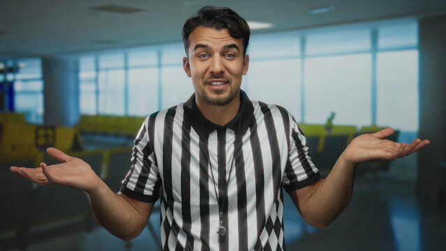 Young hispanic man dressed as referee gestures in an airport terminal conveying uncertainty and humor with hands up in an indoor setting.