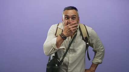 Fototapeta premium Middle age man with grey hair and beard wearing a backpack with hand covering mouth gesture in studio with purple backdrop; surprise adventure travel.
