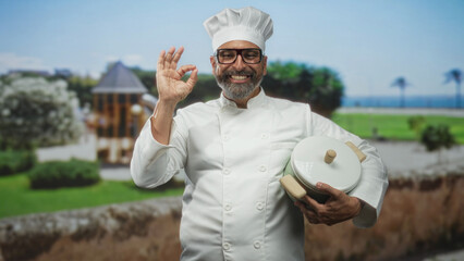 Man holding pot and making ok gesture in park with green lawn and trees visible smiling; culinary pride.