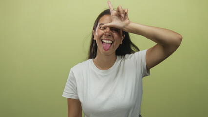Woman showing tongue out and hand forming l sign to forehead in green studio; playful rebellious vibrant energy.