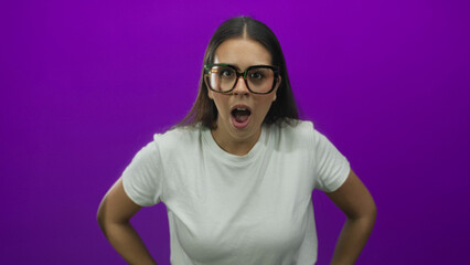 Young brunette woman wearing oversized glasses with mouth open gasping and hands on hips in studio with purple backdrop; surprise curiosity.