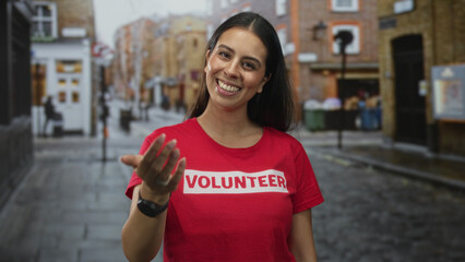 Woman volunteer in red t shirt holds out open palm in a city street, smiling and gesturing with...