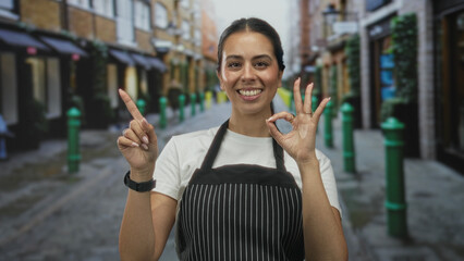 Woman in striped apron making ok sign and pointing index finger toward camera on a cobbled street...