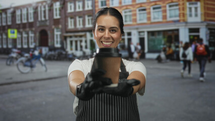 Woman in apron holding a paper cup on street in front of a cafe storefront, smiling and offering...