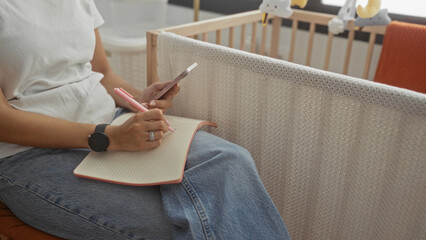 Woman writing with pen and holding smartphone by a crib in a nursery, hands on notebook across denim lap, seated indoors; calm planning motherhood.