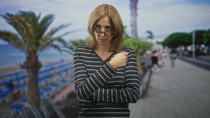 Fototapeta premium Blonde woman wearing glasses with arms crossed, peering over lenses on a seaside promenade lined with palm trees and beach chairs; disapproval judgment.
