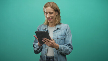 Woman holding tablet tapping screen with hands visible, smiling and standing in studio teal backdrop; calm focus.