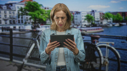 Woman holding tablet with both hands and tapping with thumbs, wearing denim jacket beside a bicycle on a street by a canal and row of buildings  concentration. © Krakenimages.com