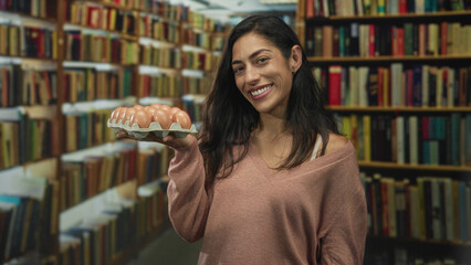 Woman holds egg carton with eyes closed and smiling while tilting head among bookshelves in a library building  curiosity domestic. © Krakenimages.com