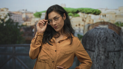 Woman smiling and adjusting glasses with hand on hip at roman building terrace overlooking ruins;...