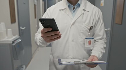 Front view close up shot of professional male doctor holding smartphone in hand and wearing lab coat while walking towards camera in clinic hall, copy space - Powered by Adobe