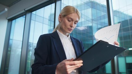 Focused Caucasian woman holding folder and examining financial charts near window inside office building. Analyzing performance results or tracking progress. Comparing data and making notes. - Powered by Adobe