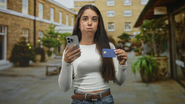 Woman holding smartphone and creditcard, puffing cheeks while showing card in a pedestrian street; happiness trust convenience. - Powered by Adobe