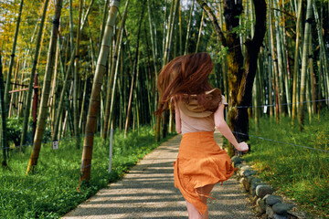 Obraz premium Woman running along a winding bamboo forest path in summer, orange skirt flowing, back view showing motion and carefree movement among tall green stalks on a shaded trail.