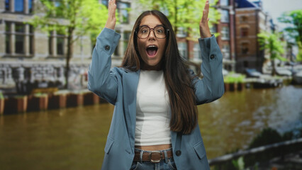 Fototapeta premium Young hispanic woman with hands raised and open mouth in surprise, wearing glasses and a blazer, standing on a street beside a canal; surprise travel.