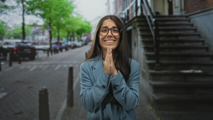 Fototapeta premium Young woman in glasses crossing both fingers with hands raised and smiling on a city street beside stone stairs and parked cars; hope gratitude.
