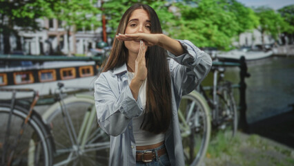 Obraz premium Young woman with long hair forms time out gesture with hands near a parked bicycle and canal set in a studio; confidence.