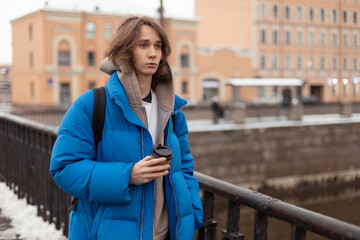 A young man walks down the street in winter wearing a hoodie and a down jacket, holding a takeaway coffee in his hand.