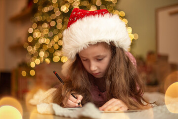 A child in a red Santa hat writes a letter or wish list next to the Christmas tree