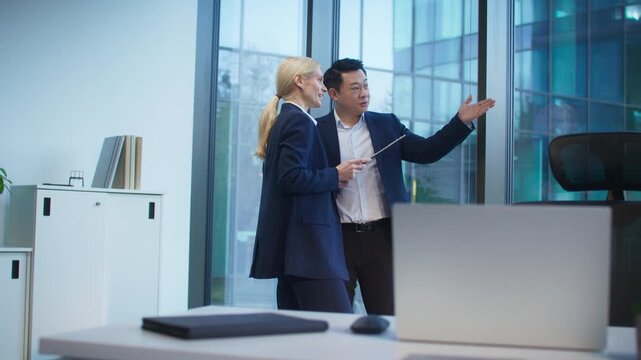 Positive Caucasian female gesturing while discussing with confident male colleague near window. Holding clipboard. Exchanging ideas or giving feedback. Planning next business steps together.