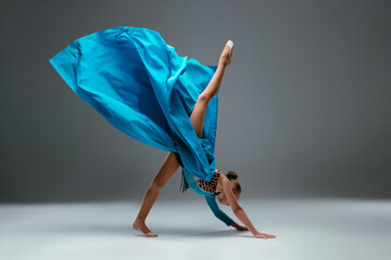 Dancer in sport leotard doing Split. Cheerleader dancing with flying blue cloth fabric. Graceful ballerina dancing on grey studio background. Child, artist, movement, action and motion concept