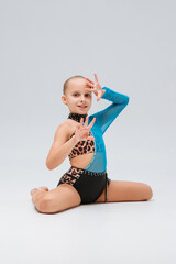 A gymnast in a striking blue and leopard-print costume, adorned with rhinestones, sits elegantly on the floor. Her raised arms emphasize poise, flexibility, and grace.