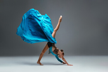 Graceful ballerina dancing on grey studio background. Cheerleader dancing with flying blue cloth fabric. Dancer in sport leotard doing Split. Child, artist, movement, action and motion concept