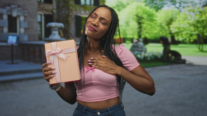 Black woman holds a pink gift box with ribbon, hand pressed to chest gesture, wearing pink shirt...