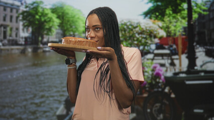Fototapeta premium Black woman holding a cake on a street canal, eyes closed and smelling pastry with hands supporting wooden board; culinary contentment.